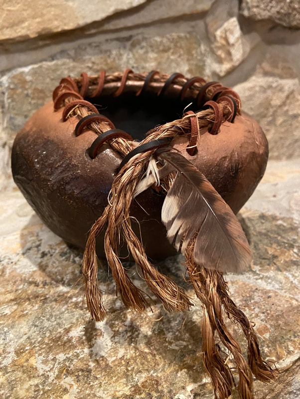 Small mica clay pot with texture. Around the top are holes used to lace rope and leather. A feather is intertwined in the rope and leathers. Burnished and pit fired. 